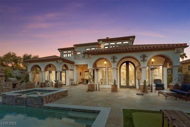 back house at dusk featuring french doors, a balcony, a pool with hot tub, and a patio