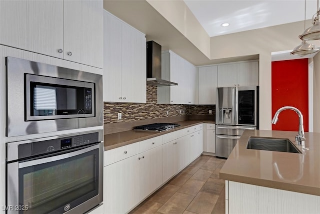 kitchen with appliances with stainless steel finishes, white cabinetry, sink, hanging light fixtures, and wall chimney exhaust hood