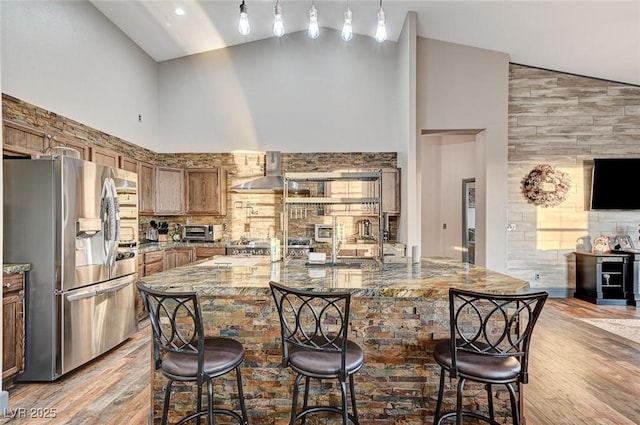 kitchen with wall chimney exhaust hood, light stone counters, high vaulted ceiling, light hardwood / wood-style flooring, and stainless steel fridge