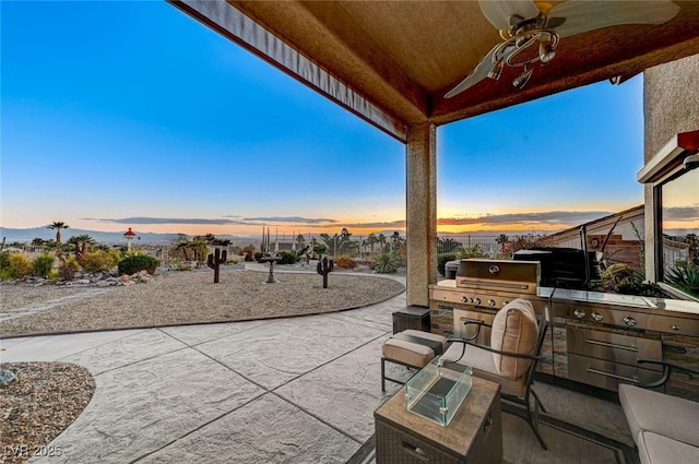 patio terrace at dusk featuring an outdoor kitchen, ceiling fan, and area for grilling