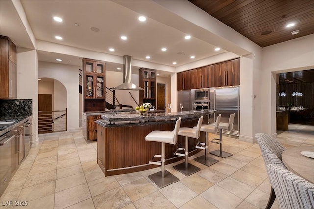 kitchen featuring tasteful backsplash, built in appliances, a breakfast bar area, a large island, and wall chimney exhaust hood