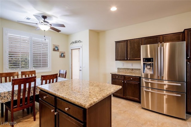 kitchen with stainless steel fridge, ceiling fan, dark brown cabinets, light stone countertops, and a kitchen island