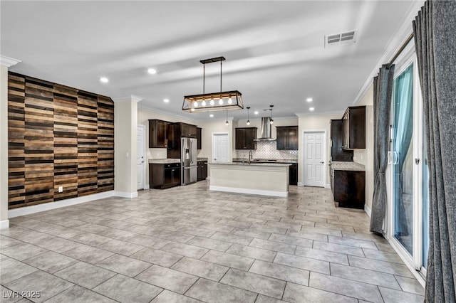 kitchen with an island with sink, decorative backsplash, stainless steel fridge with ice dispenser, crown molding, and wall chimney range hood