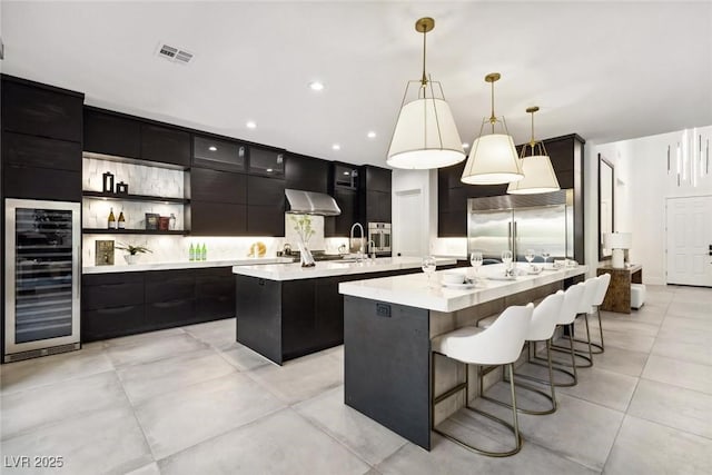 kitchen featuring a kitchen island with sink, stainless steel appliances, wine cooler, decorative light fixtures, and wall chimney exhaust hood