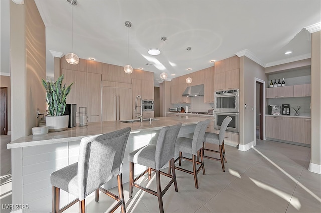 kitchen with sink, wall chimney exhaust hood, decorative light fixtures, stainless steel double oven, and light brown cabinets