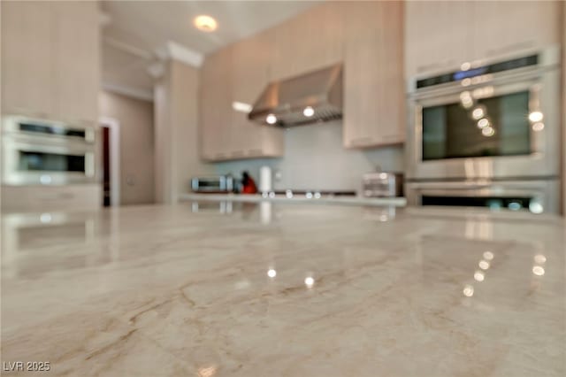 kitchen with light brown cabinetry, ventilation hood, double oven, and light stone countertops