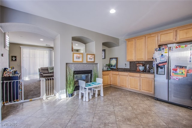 kitchen with light brown cabinetry, light tile patterned floors, stainless steel fridge, dark stone counters, and a tiled fireplace