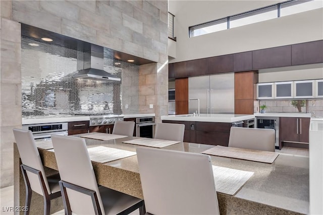 kitchen with wall chimney exhaust hood, dark brown cabinetry, appliances with stainless steel finishes, beverage cooler, and backsplash