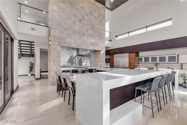 kitchen featuring wall chimney range hood, built in refrigerator, backsplash, a spacious island, and a kitchen bar