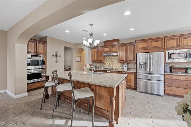 kitchen with appliances with stainless steel finishes, an island with sink, sink, hanging light fixtures, and light stone counters