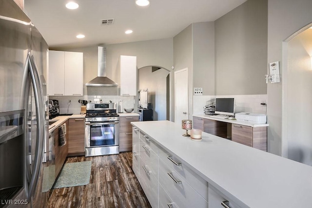 kitchen with appliances with stainless steel finishes, dark hardwood / wood-style flooring, wall chimney range hood, decorative backsplash, and white cabinets