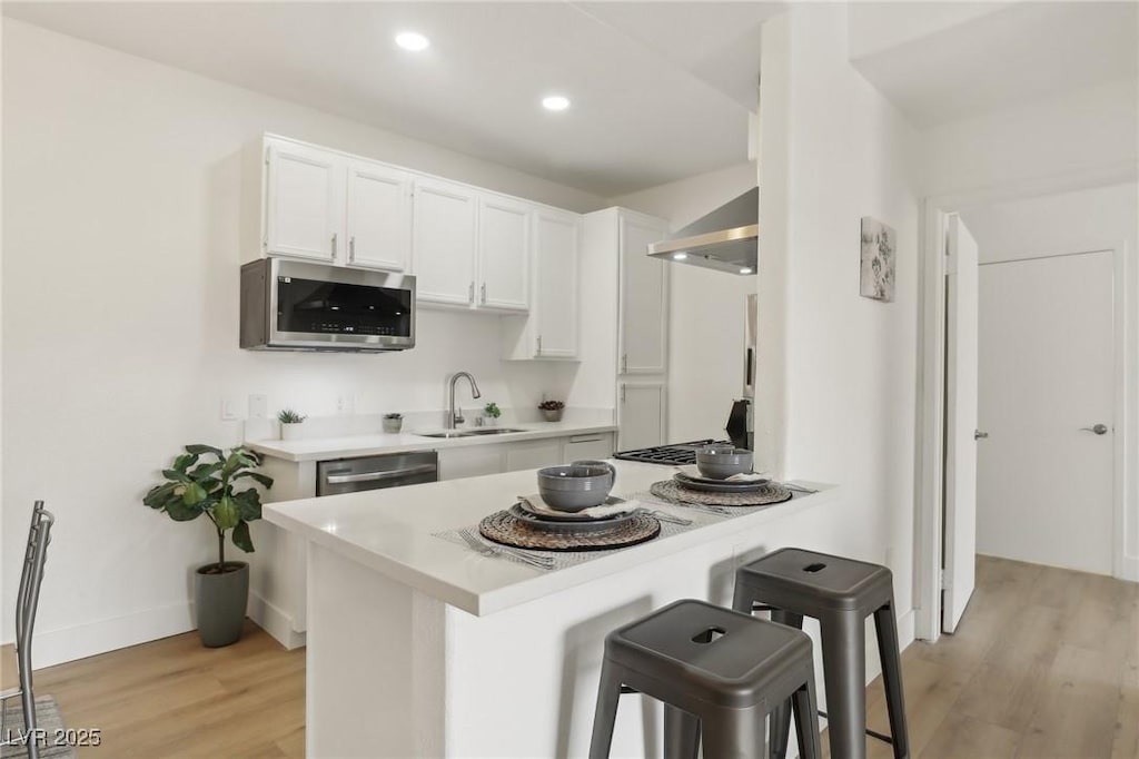 kitchen with a breakfast bar, sink, appliances with stainless steel finishes, white cabinets, and wall chimney range hood