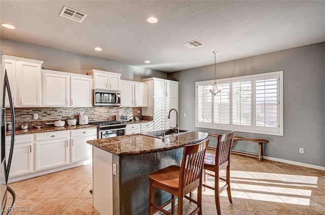 kitchen with sink, appliances with stainless steel finishes, dark stone counters, a kitchen island with sink, and white cabinets