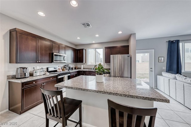 kitchen with a breakfast bar area, appliances with stainless steel finishes, dark brown cabinets, light stone countertops, and light tile patterned flooring