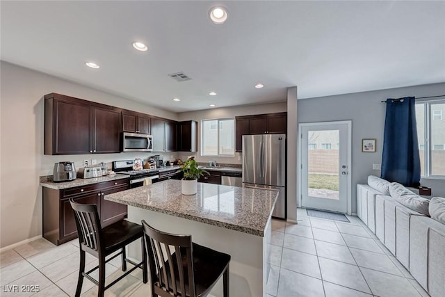 kitchen with light tile patterned flooring, a kitchen island, appliances with stainless steel finishes, a kitchen bar, and light stone counters