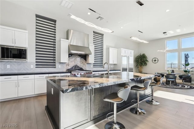 kitchen with wall chimney exhaust hood, built in appliances, decorative light fixtures, dark stone counters, and a kitchen island with sink