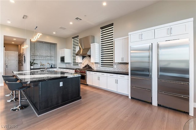 kitchen with decorative light fixtures, an island with sink, white cabinets, built in appliances, and wall chimney exhaust hood