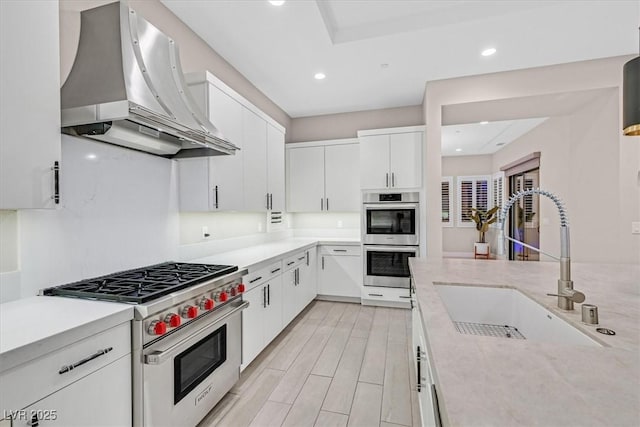 kitchen with wood tiled floor, stainless steel appliances, wall chimney range hood, white cabinetry, and a sink