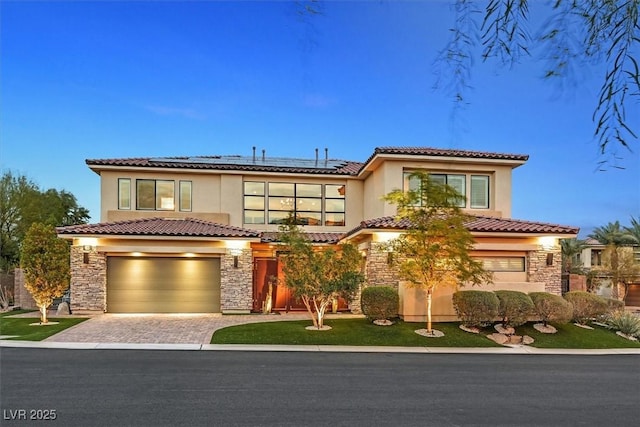 mediterranean / spanish house with a garage, stone siding, decorative driveway, roof mounted solar panels, and stucco siding