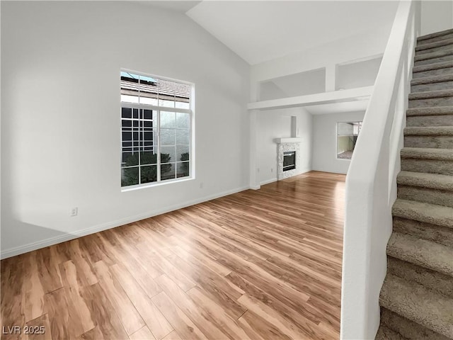 unfurnished living room featuring stairway, baseboards, light wood finished floors, vaulted ceiling, and a glass covered fireplace