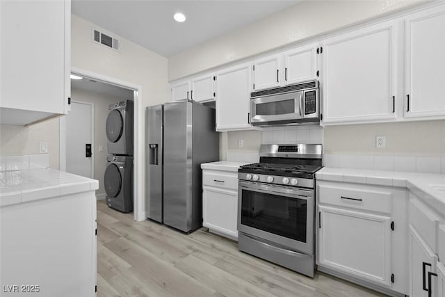 kitchen with tile countertops, white cabinets, stacked washer / drying machine, light hardwood / wood-style floors, and stainless steel appliances