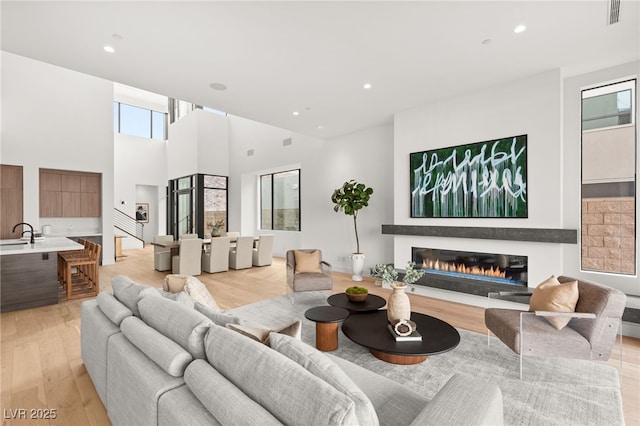 living room featuring light wood-style flooring, recessed lighting, a towering ceiling, visible vents, and a glass covered fireplace