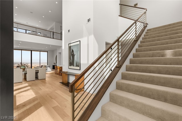 stairs featuring visible vents, a towering ceiling, wood finished floors, a wall of windows, and recessed lighting