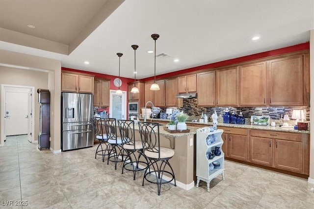 kitchen featuring a breakfast bar, stainless steel appliances, light stone countertops, an island with sink, and decorative light fixtures