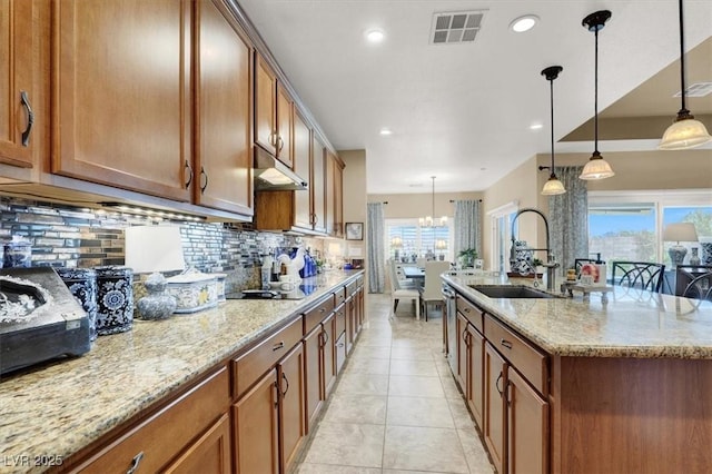 kitchen with sink, backsplash, hanging light fixtures, light stone countertops, and a center island with sink