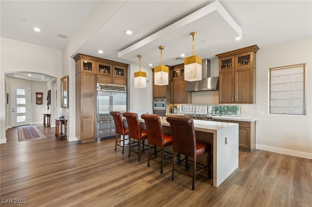 kitchen with wall chimney range hood, a breakfast bar area, appliances with stainless steel finishes, an island with sink, and decorative light fixtures