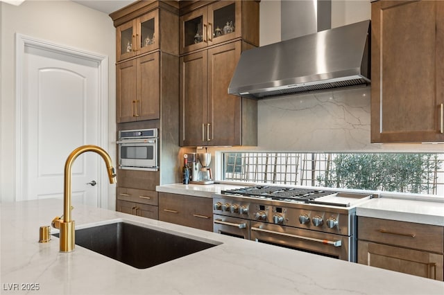 kitchen featuring wall chimney exhaust hood, sink, light stone counters, appliances with stainless steel finishes, and backsplash