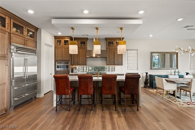 kitchen featuring pendant lighting, wall chimney range hood, stainless steel appliances, and an island with sink