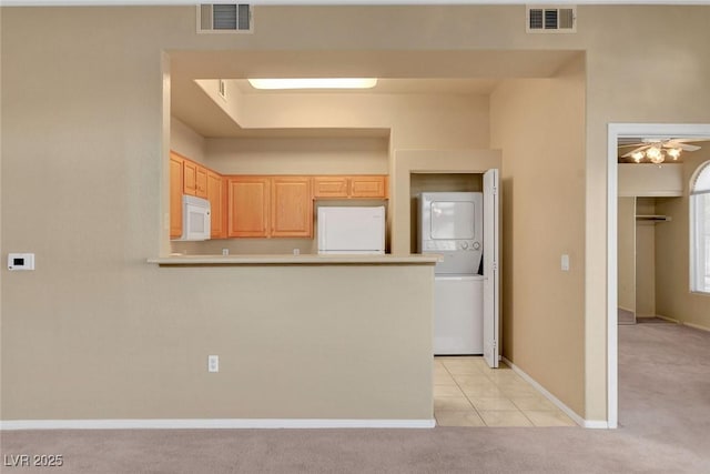 kitchen with stacked washer / drying machine, kitchen peninsula, light carpet, light brown cabinets, and white appliances