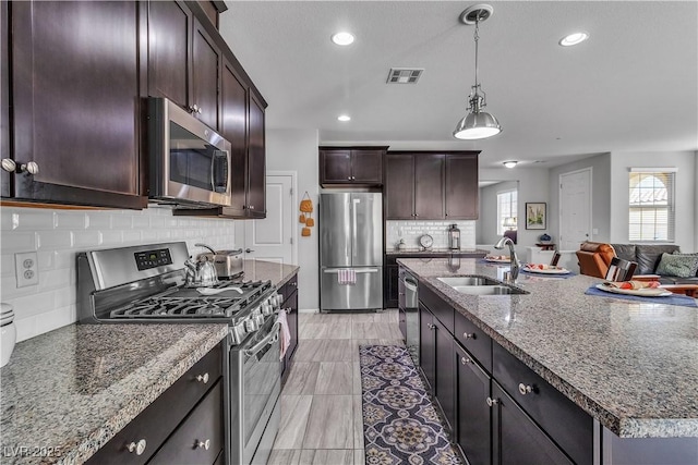 kitchen with sink, a kitchen island with sink, stainless steel appliances, dark brown cabinetry, and stone countertops