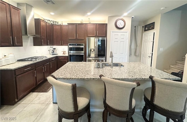 kitchen featuring wall chimney exhaust hood, a kitchen bar, sink, an island with sink, and stainless steel appliances