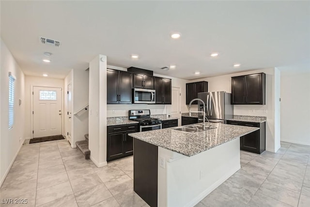 kitchen with sink, light tile patterned floors, appliances with stainless steel finishes, light stone counters, and an island with sink