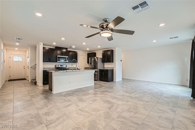 kitchen featuring sink, ceiling fan, appliances with stainless steel finishes, light stone countertops, and a center island with sink