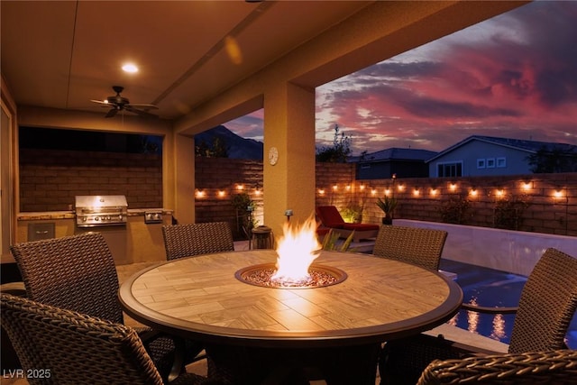 patio terrace at dusk featuring an outdoor kitchen, a grill, ceiling fan, and a fire pit