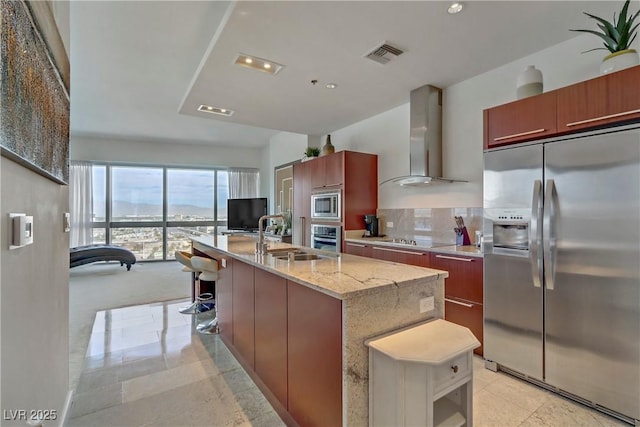 kitchen featuring sink, a center island with sink, appliances with stainless steel finishes, light stone countertops, and wall chimney range hood