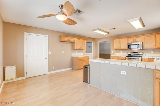 kitchen featuring tile countertops, ceiling fan, stainless steel appliances, light brown cabinets, and light hardwood / wood-style flooring