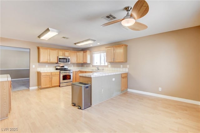 kitchen with appliances with stainless steel finishes, tile countertops, light brown cabinetry, sink, and light hardwood / wood-style floors