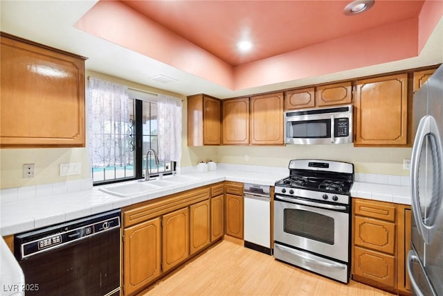 kitchen featuring appliances with stainless steel finishes, sink, tile counters, a tray ceiling, and light hardwood / wood-style flooring