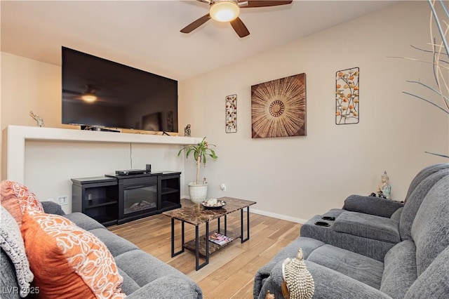 living room with light wood-type flooring, baseboards, a ceiling fan, and a glass covered fireplace