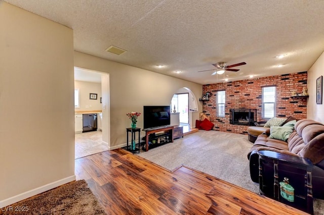 living room with ceiling fan, light hardwood / wood-style floors, a brick fireplace, and a textured ceiling