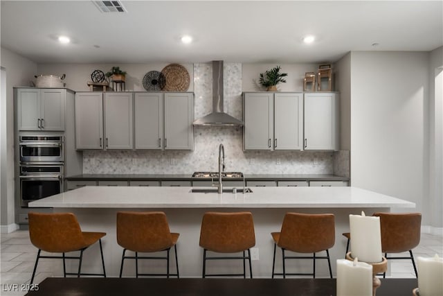 kitchen featuring gray cabinetry, stainless steel appliances, tasteful backsplash, an island with sink, and wall chimney exhaust hood