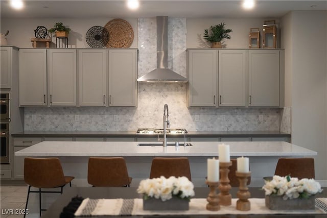 kitchen with gray cabinets, tasteful backsplash, an island with sink, stainless steel appliances, and wall chimney exhaust hood