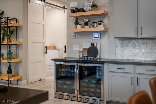 bar featuring gray cabinetry, a barn door, light tile patterned floors, beverage cooler, and decorative backsplash