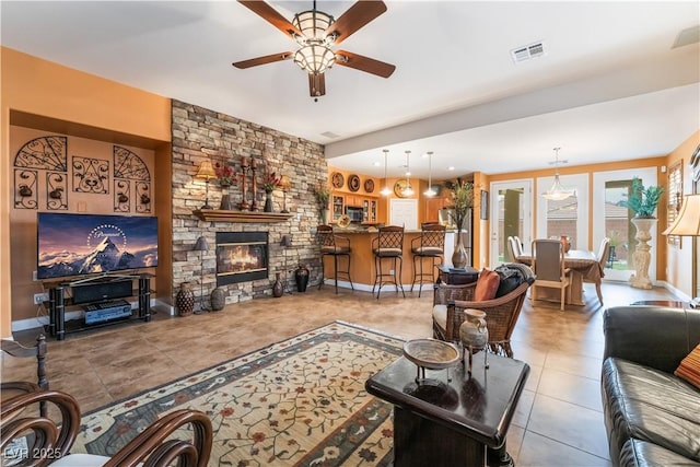 living area with visible vents, light tile patterned flooring, ceiling fan, a stone fireplace, and baseboards