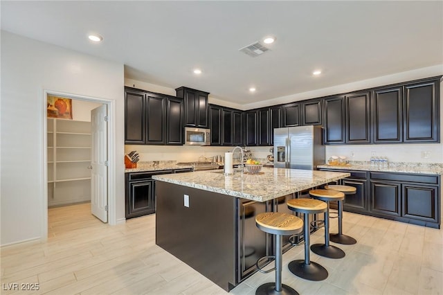 kitchen with appliances with stainless steel finishes, a breakfast bar, a kitchen island with sink, light hardwood / wood-style floors, and light stone countertops