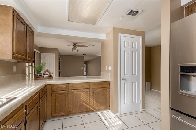 kitchen with light tile patterned floors, tile counters, stainless steel fridge, kitchen peninsula, and ceiling fan